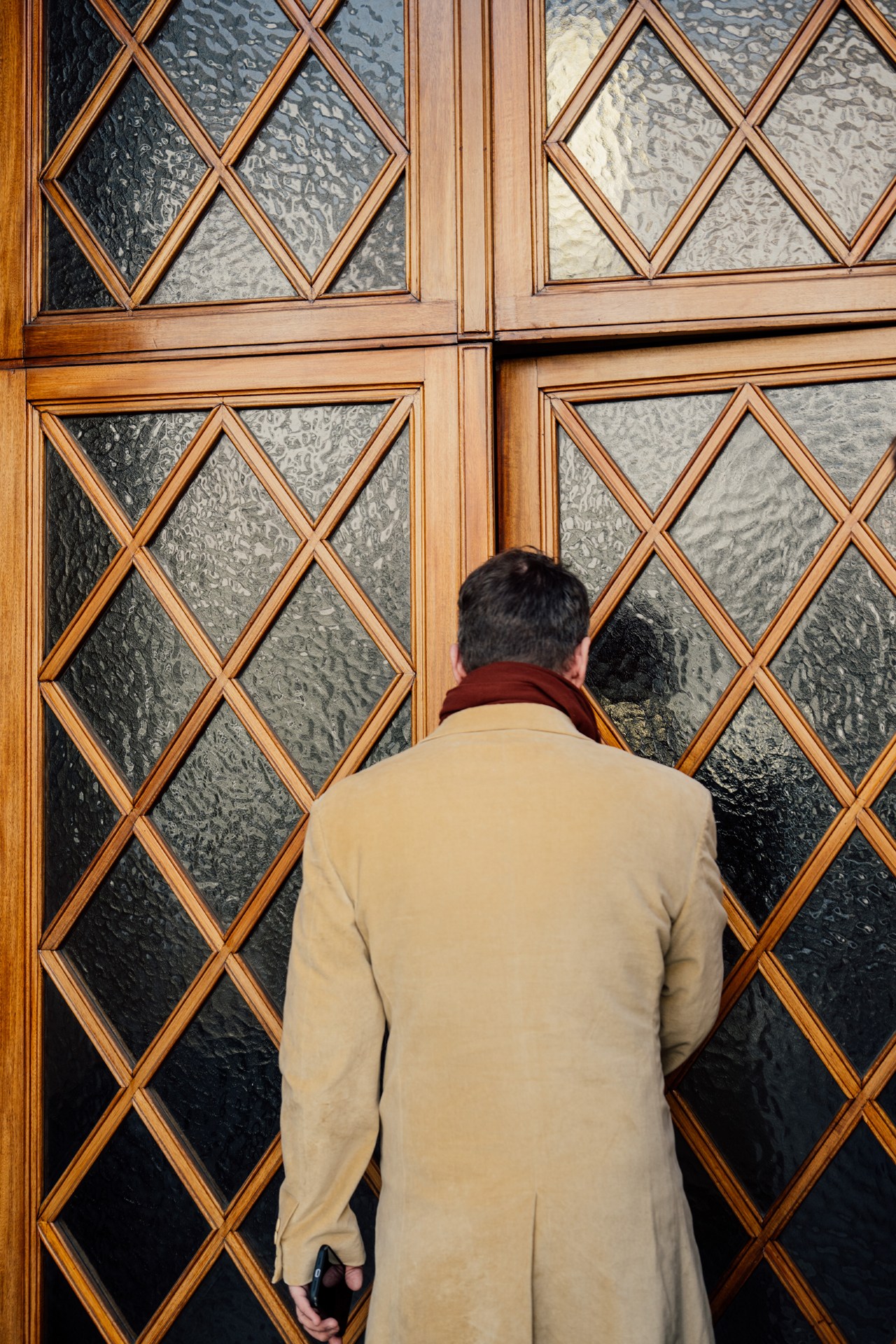 Person in beige coat examining ornate glass-paneled wooden doors with diamond lattice pattern