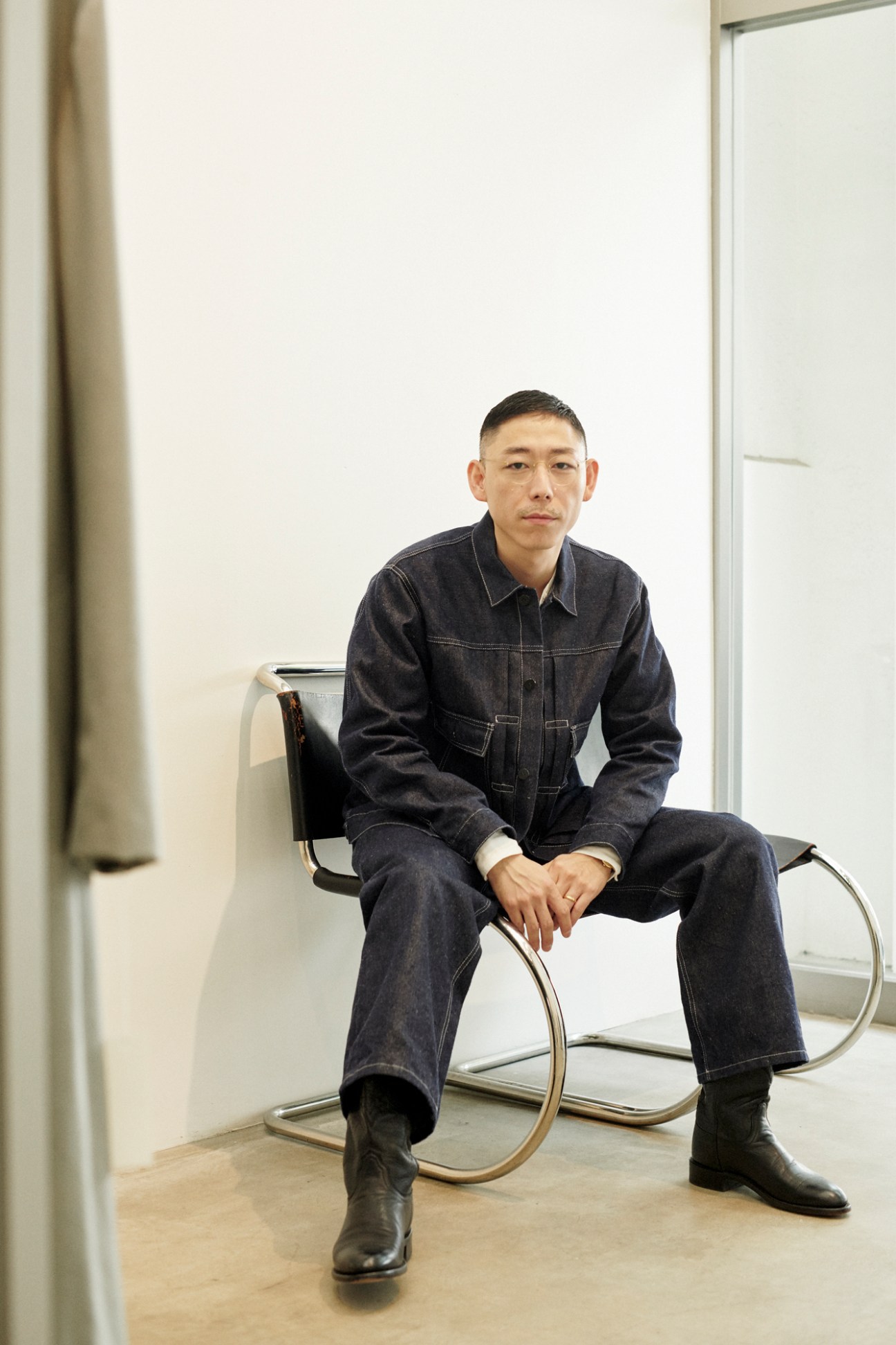 Yuki Sato sitting in modern chair wearing dark denim outfit in minimalist white interior space