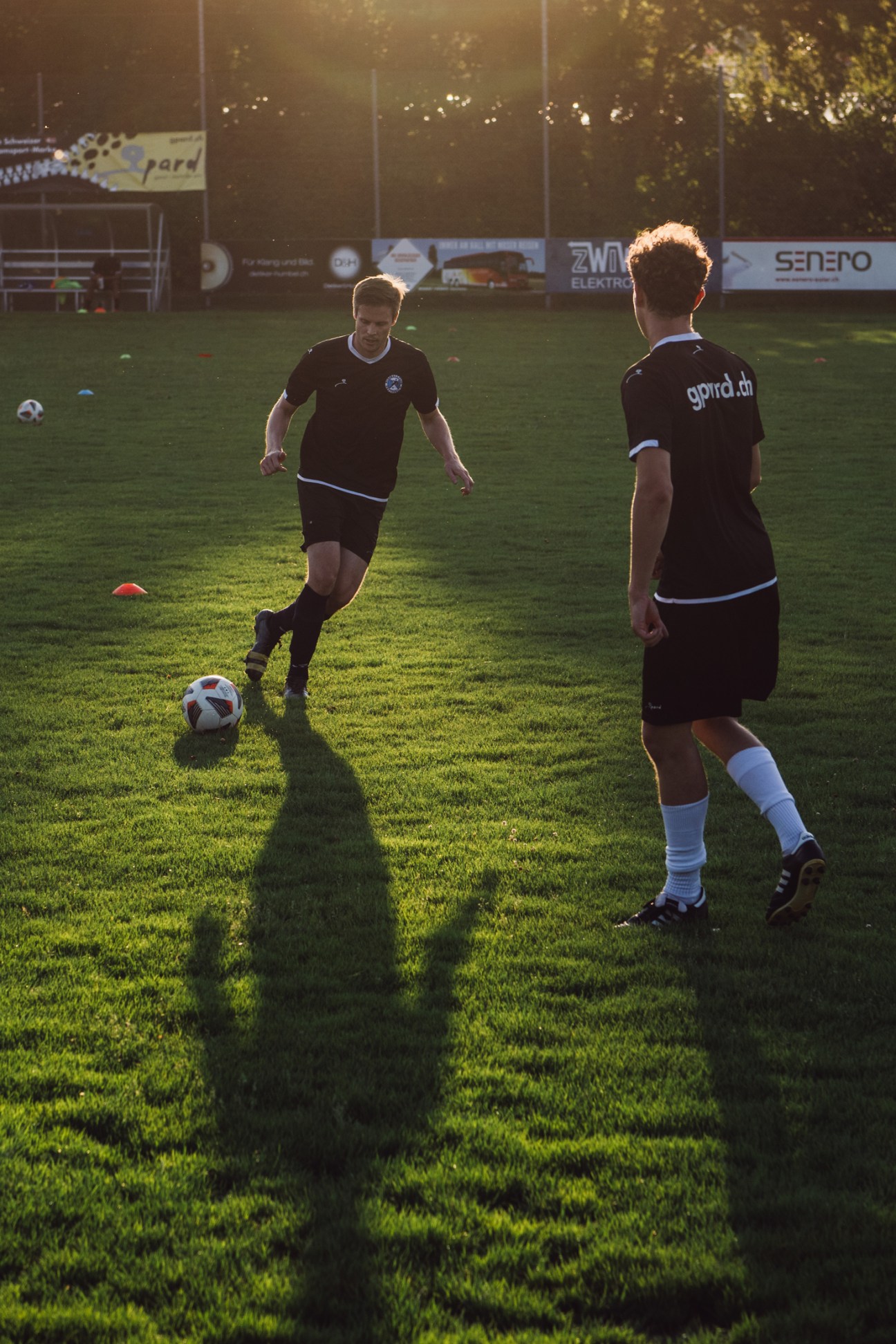 Two soccer players in dark uniforms practicing on a grass field during golden hour with stadium signage visible