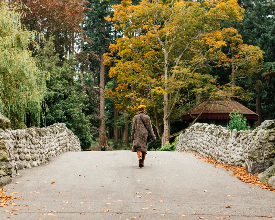 Person in brown coat walking alone on autumn path lined with stone walls.