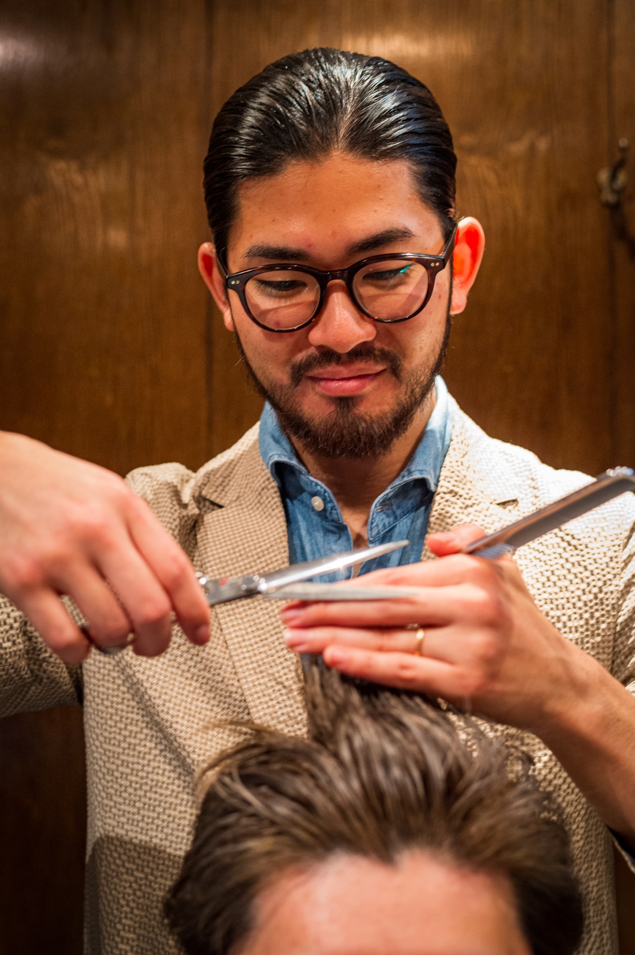 A barber wearing glasses and a beige cardigan carefully cuts a client's hair with scissors in a wooden-paneled salon