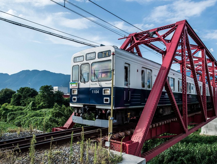 A two-tone train crossing a red metal bridge with mountains visible in the background.