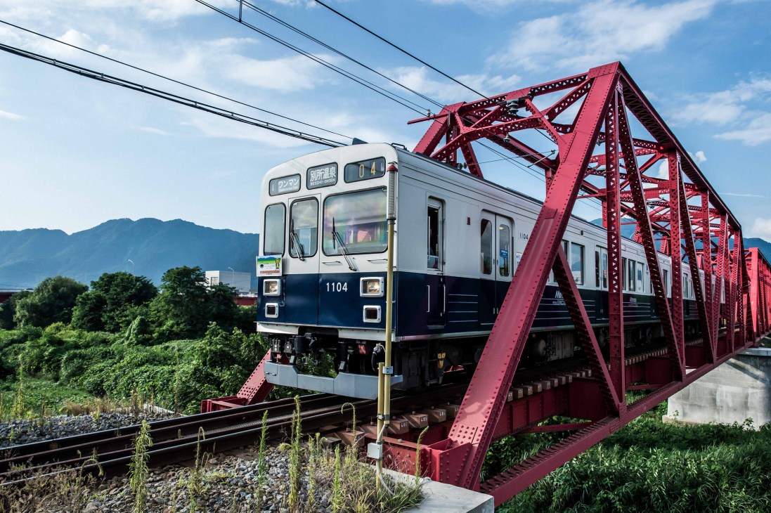 A two-tone train crossing a red metal bridge with mountains visible in the background.
