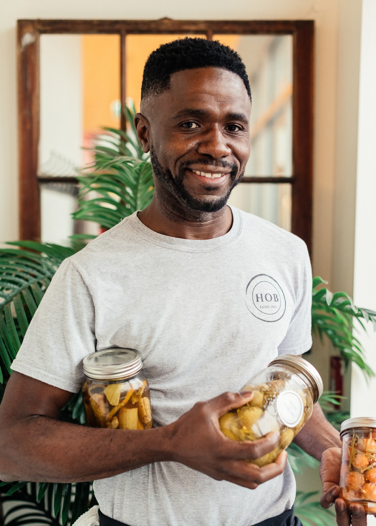 Man in HOB Food Inc t-shirt holding glass jars of preserved vegetables in bright interior.