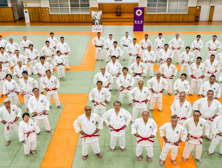 Large group of judo practitioners in white gis with red belts positioned across green and orange tatami mats in dojo