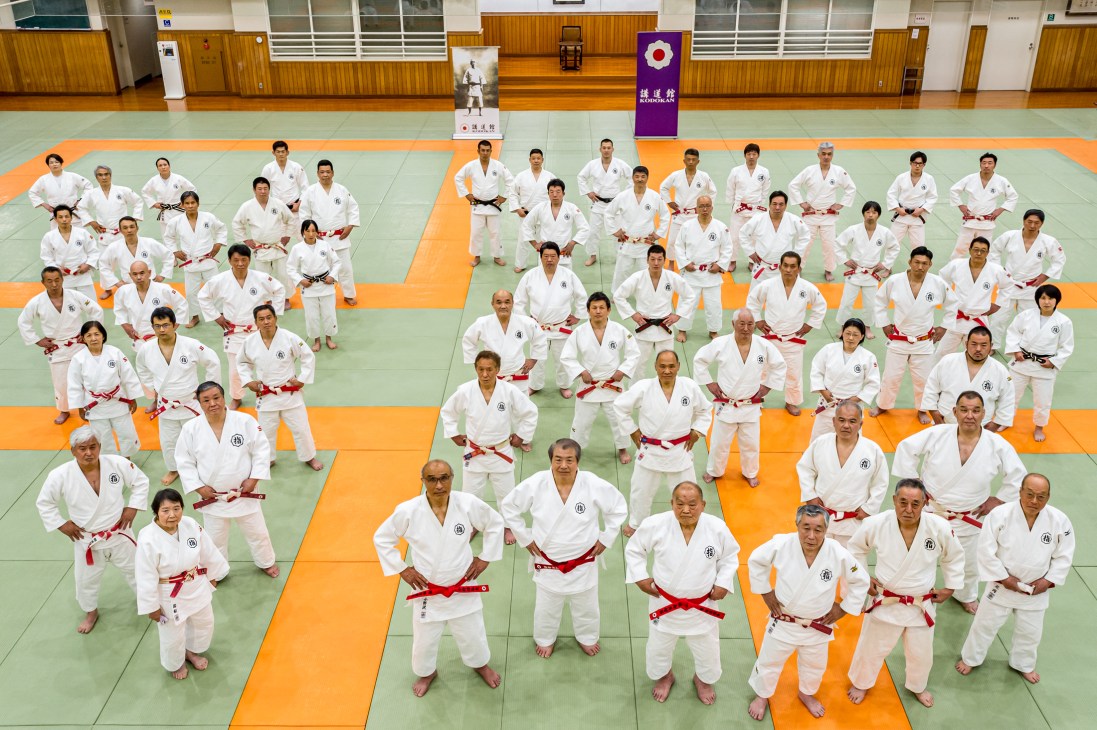 Large group of judo practitioners in white gis with red belts positioned across green and orange tatami mats in dojo
