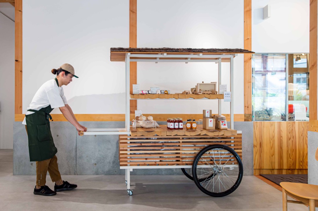 Person in white shirt and cap pushing wooden mobile cart with shelved products and bicycle wheel