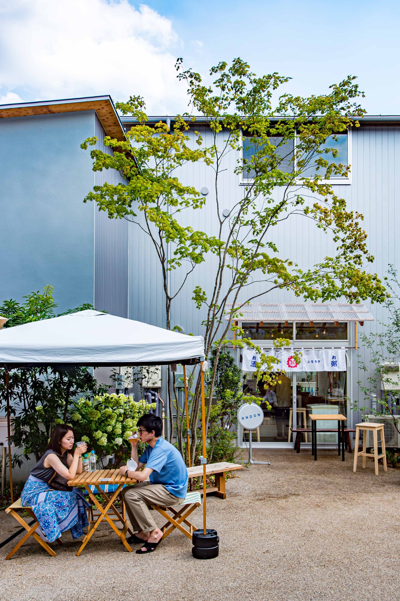 Two people sitting at outdoor table under umbrella at modern cafe with corrugated metal building in background