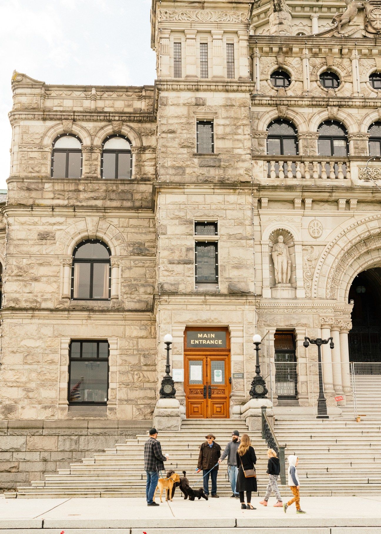 People walking on stone steps outside a historic neoclassical building with ornate architectural details and an orange wooden main entrance door.