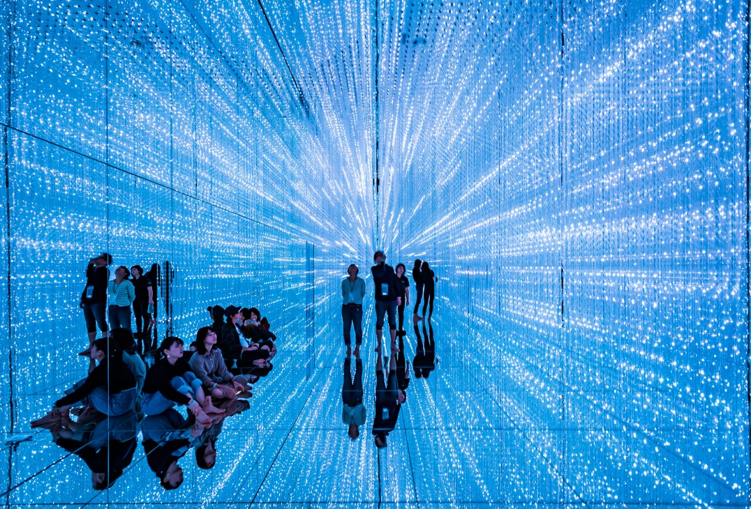 Visitors silhouetted against blue digital display with radiating light patterns in TeamLab's Infinite Crystal Universe installation.