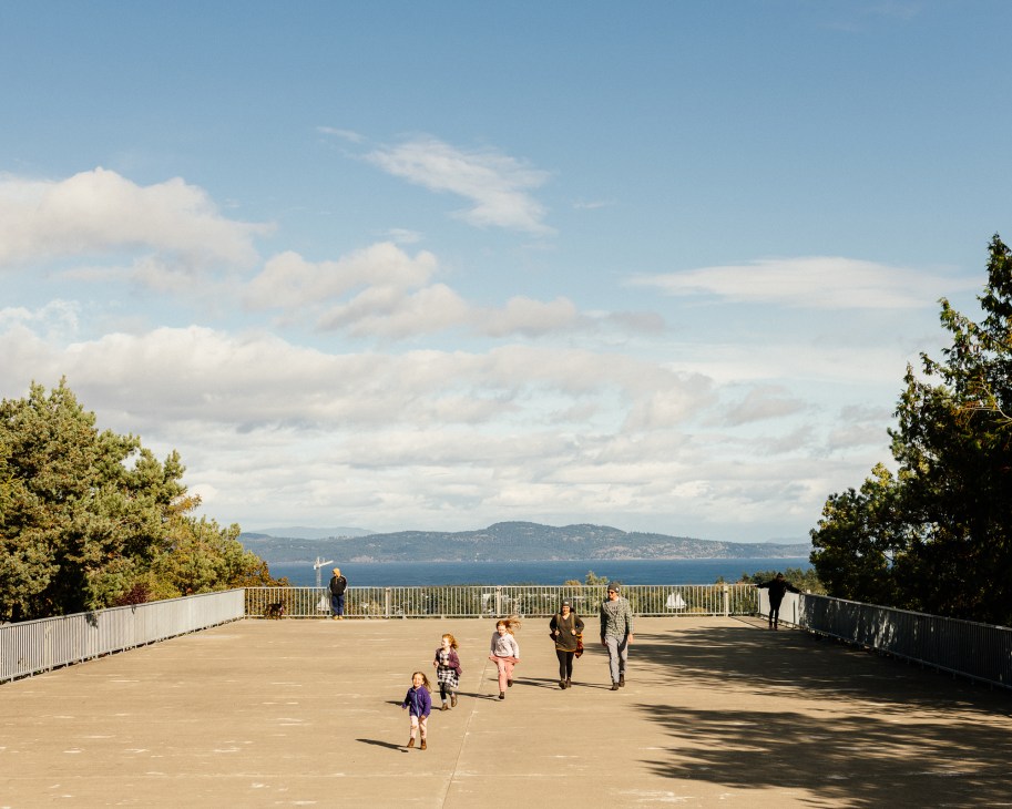People walking and cycling on a sunny wooden boardwalk overlooking Victoria, Canada's waterfront and distant mountains.