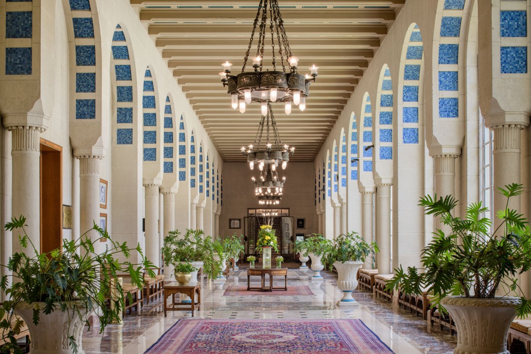Elegant colonnade hallway with white arches, blue tile details, ornate chandeliers and potted plants