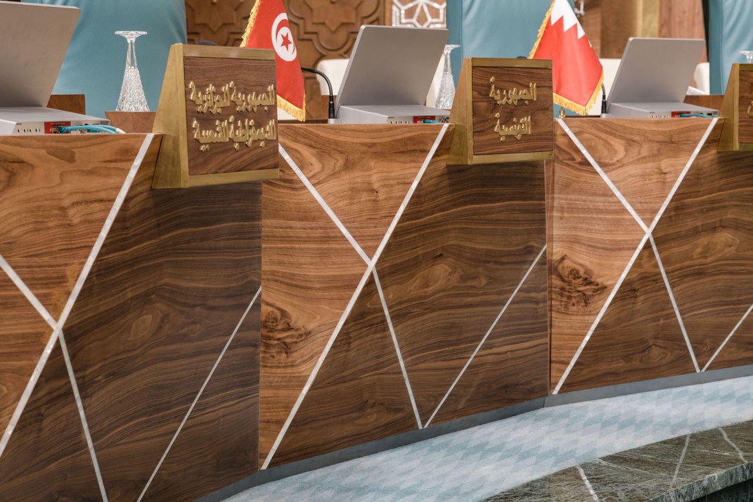 Wooden desks with geometric white mother-of-pearl inlay patterns and Arabic nameplates, with flags in background