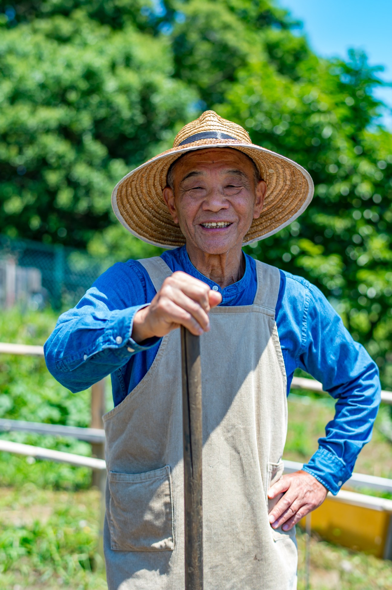 Kichio Kimura, an elderly man in traditional farming attire with a wide-brimmed hat, smiles while holding a garden tool