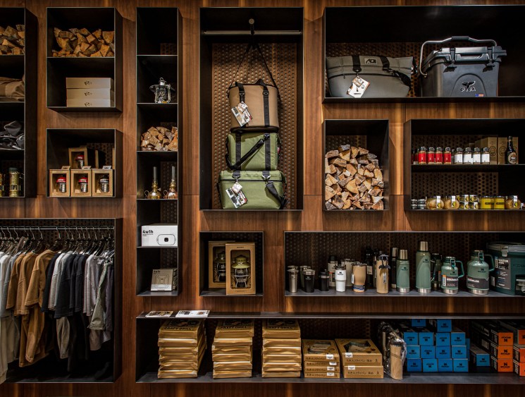 Wooden display wall shelving unit featuring organized retail merchandise including hanging clothing in earth tones on the left, stacked outdoor gear and camping equipment, firewood bundles, bottled products, coolers, thermos containers, and packaged goods in blue and orange boxes on lower shelves.