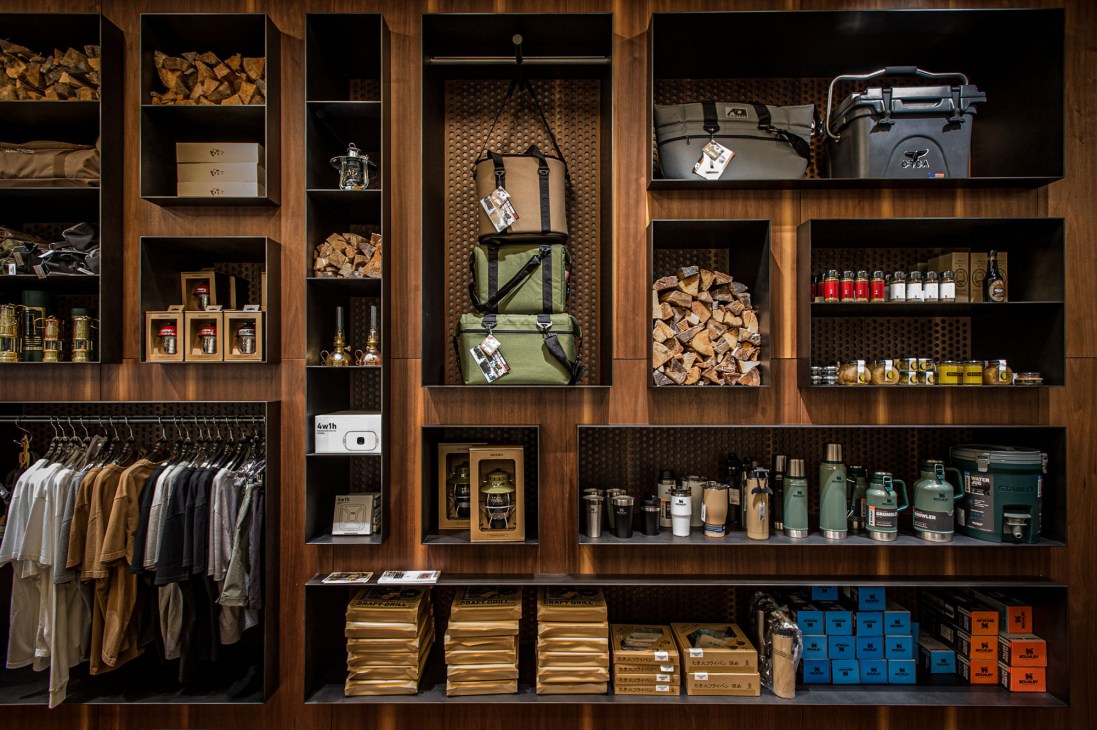Wooden display wall shelving unit featuring organized retail merchandise including hanging clothing in earth tones on the left, stacked outdoor gear and camping equipment, firewood bundles, bottled products, coolers, thermos containers, and packaged goods in blue and orange boxes on lower shelves.