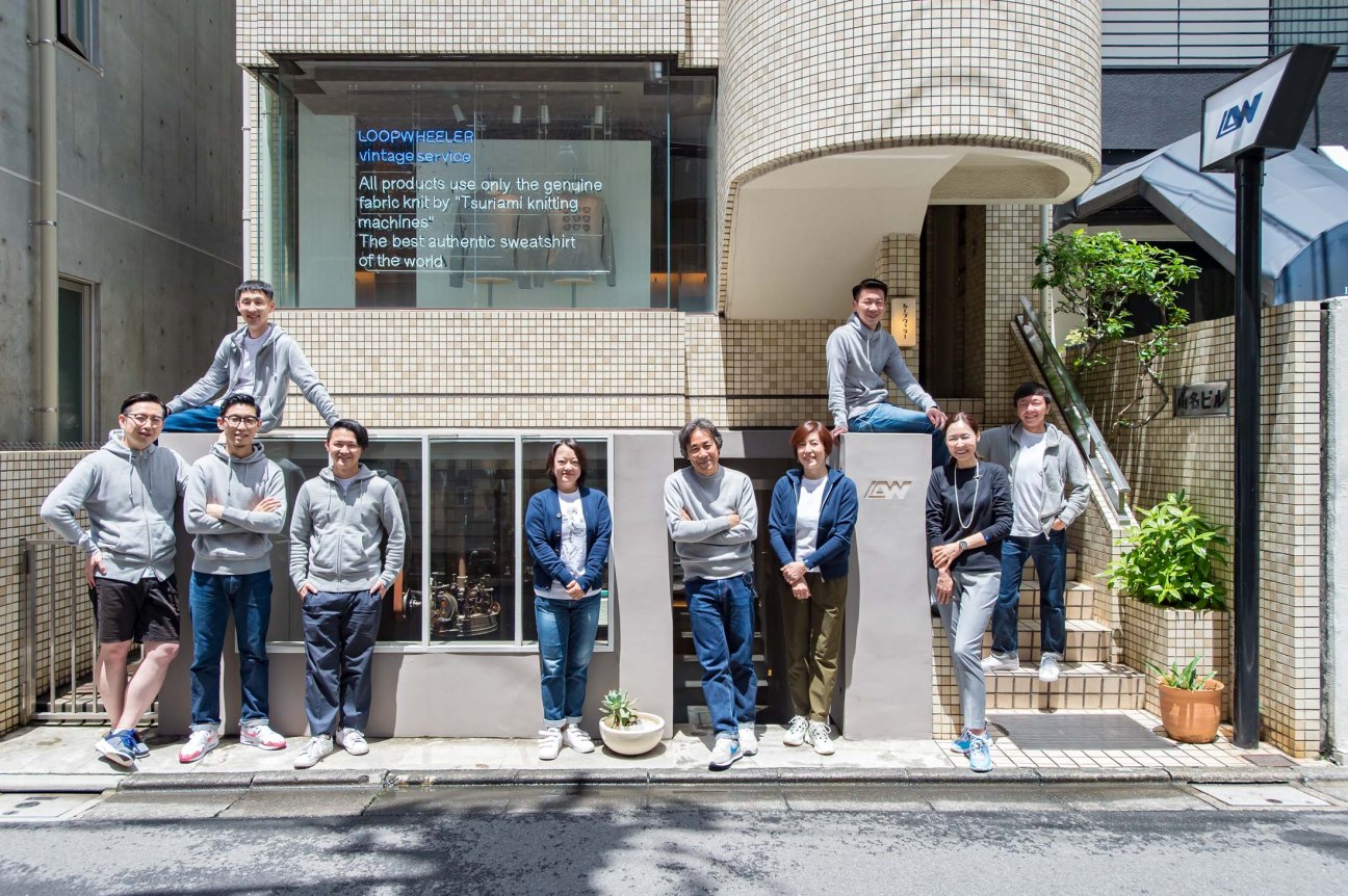 Team of Loopwheeler staff posed outside their Tokyo storefront with signage about their Tsunami knitting machines.