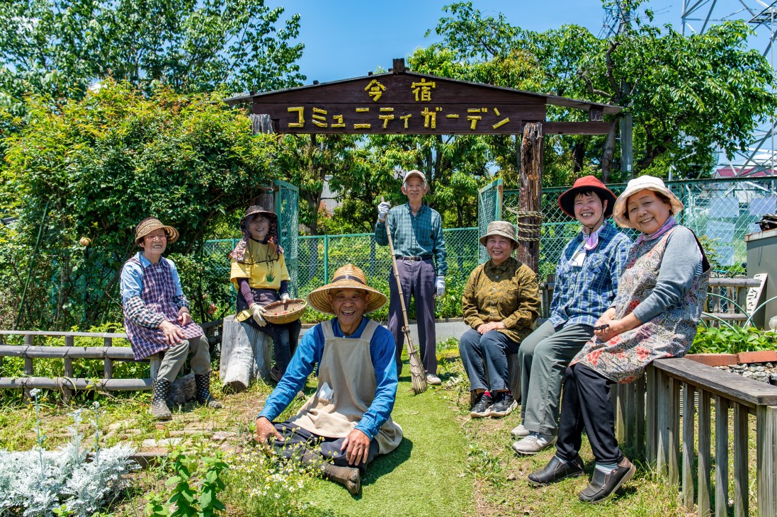 Group of seven elderly Japanese gardeners in hats and work clothes posing together at Imajuku Community Garden