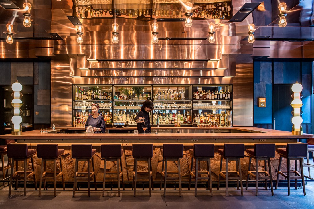 Modern bar interior with copper walls, exposed ceiling, string lights, and black bar stools facing bartenders