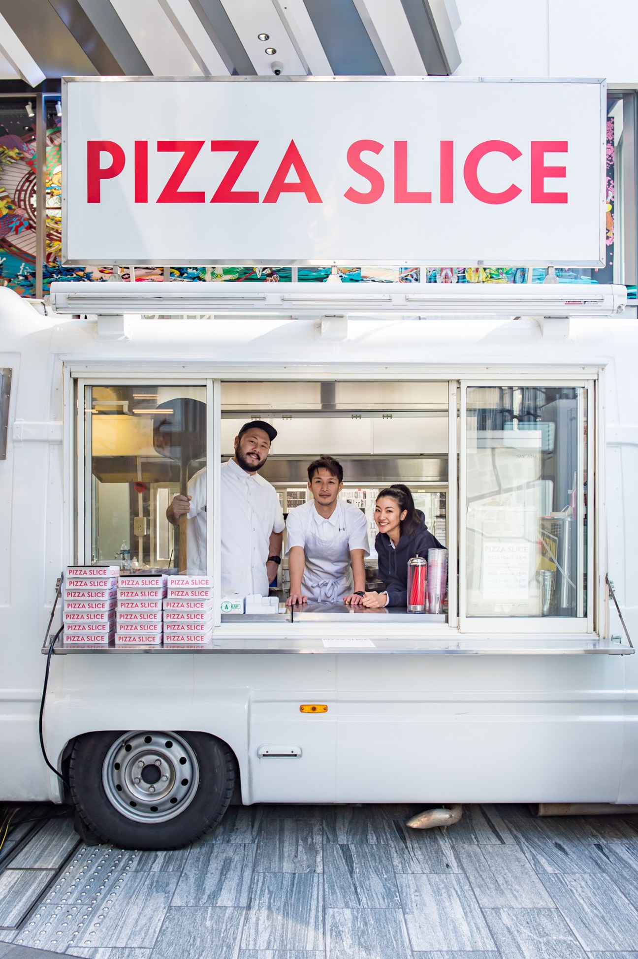 Three people standing in service window of white pizza food truck with Pizza Slice signage