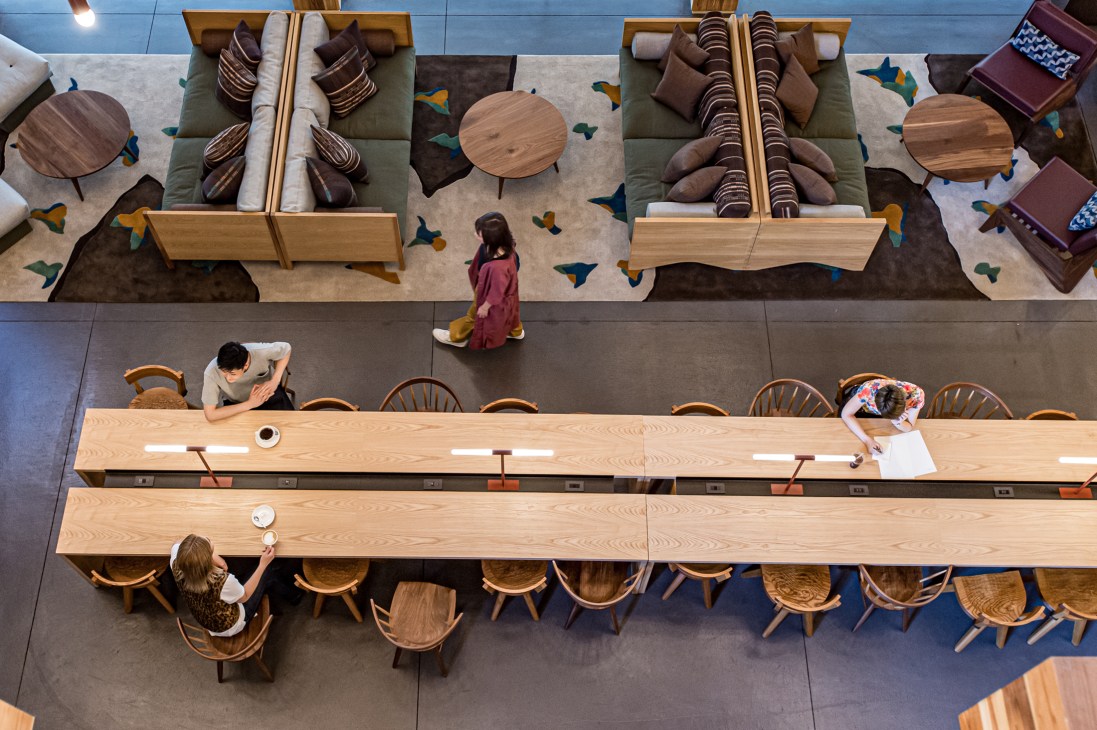 Aerial view of hotel lobby workspace with long wooden communal table, chairs, and lounge seating areas