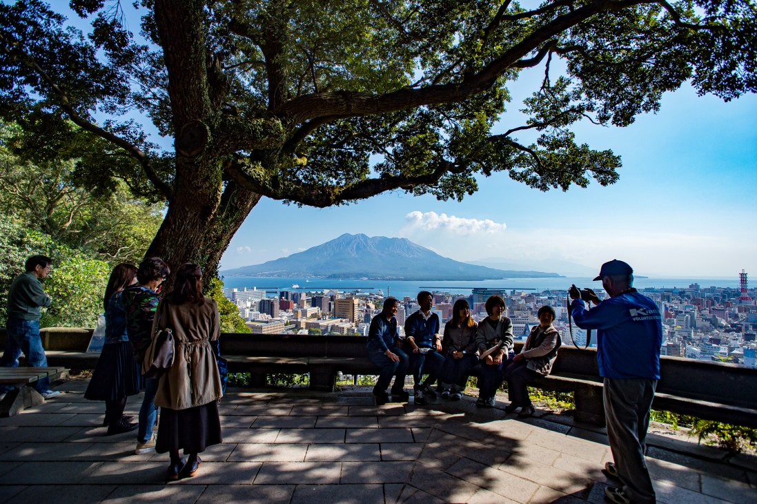 Tourists sitting on a bench under a tree overlooking Kagoshima city with Sakurajima volcano in the distance
