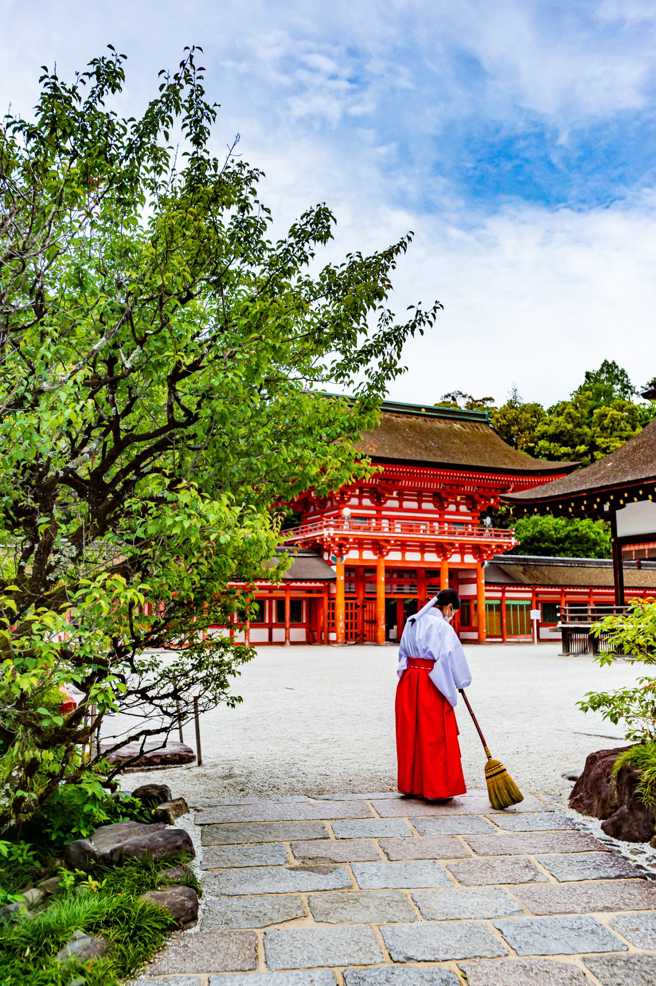 Person in traditional white and red clothing sweeping the courtyard of a red Japanese temple with green trees overhead