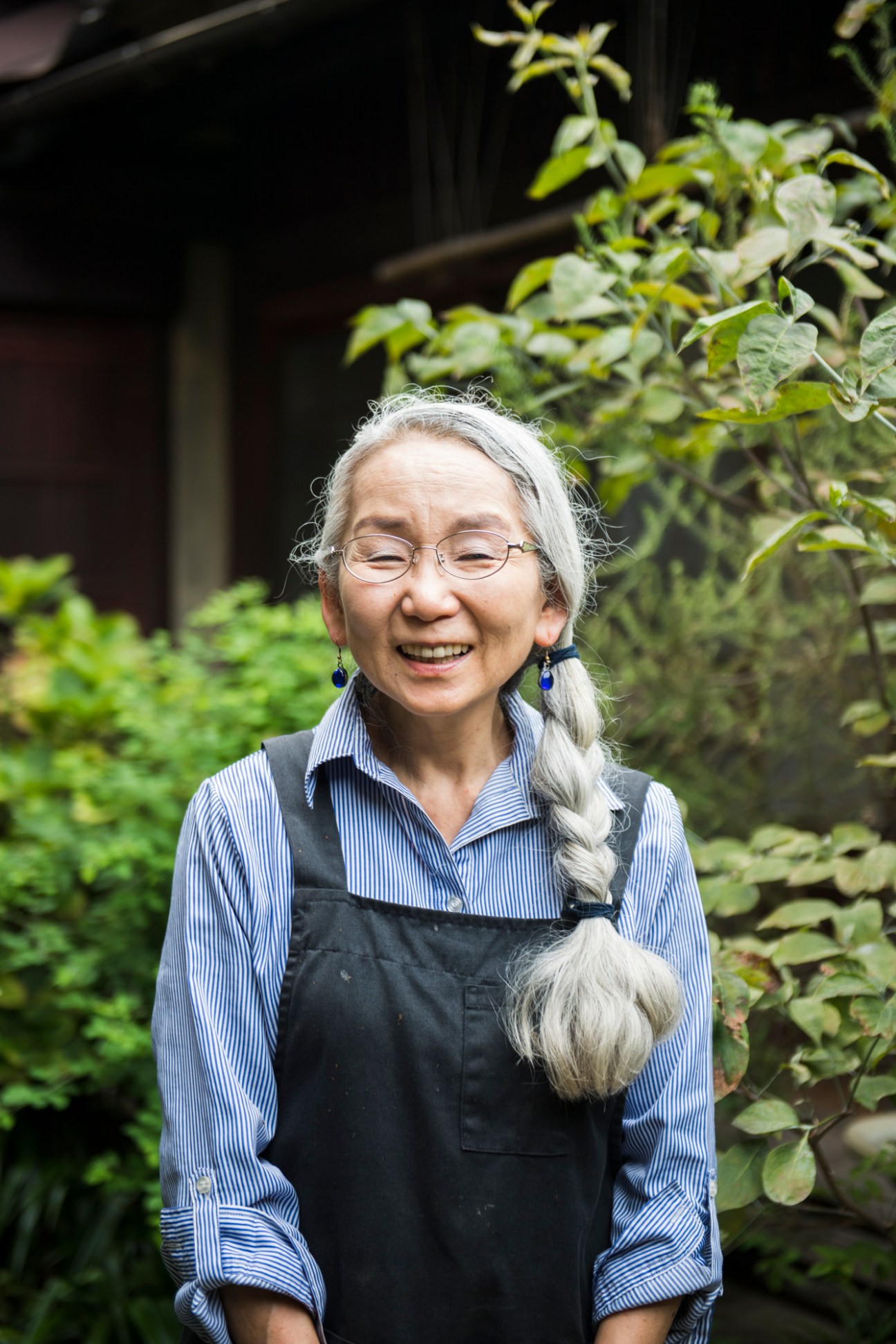 Kaneko Kikkawa smiling in a garden wearing glasses, a striped shirt and black apron at Sowakarou Inn in Tomo, Japan.