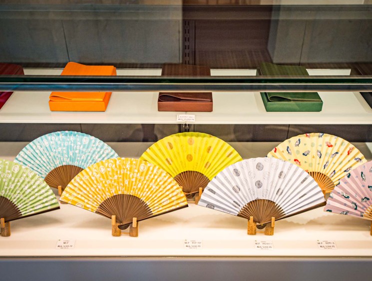 Colorful traditional Japanese folding fans with wooden handles displayed on white shelving with decorative boxes above.