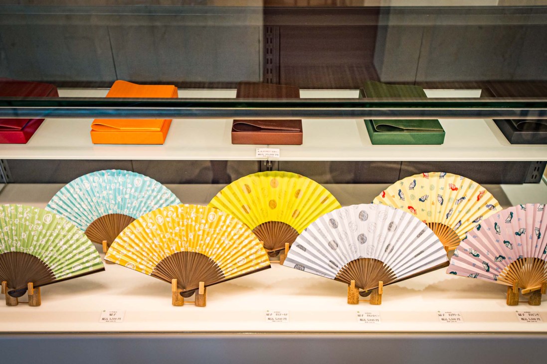 Colorful traditional Japanese folding fans with wooden handles displayed on white shelving with decorative boxes above.