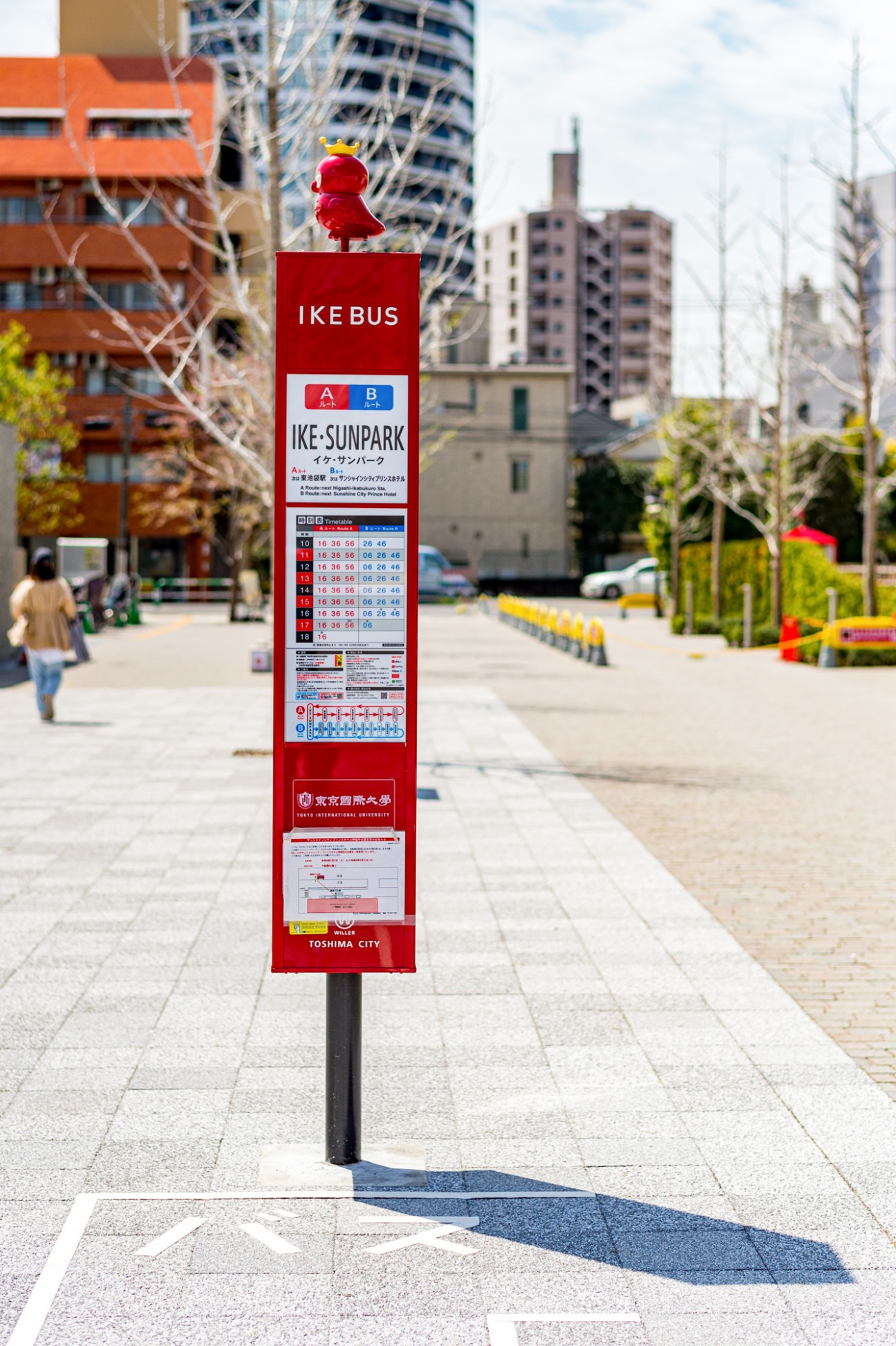 Red Ikebus stop sign with schedule and route information in urban plaza setting