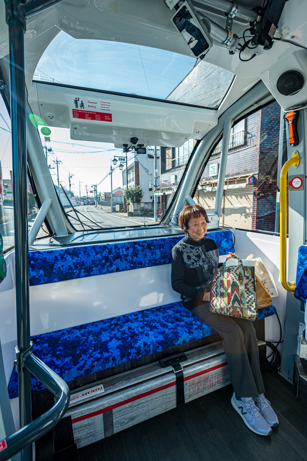 Elderly woman with shopping bag sitting on blue patterned seats inside autonomous bus interior