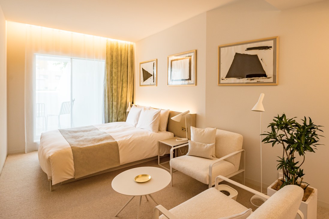 Modern hotel bedroom with neutral tones, framed artwork, seating area, and large window