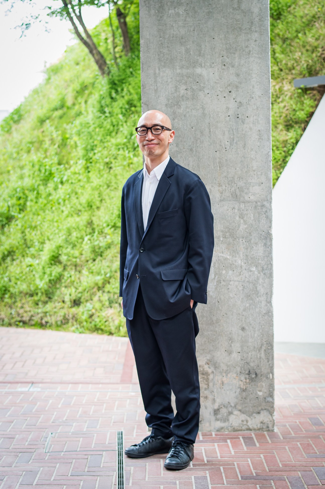 Ko Yamura, CEO of Shiroiya Hotel, standing against concrete pillar in dark suit and glasses