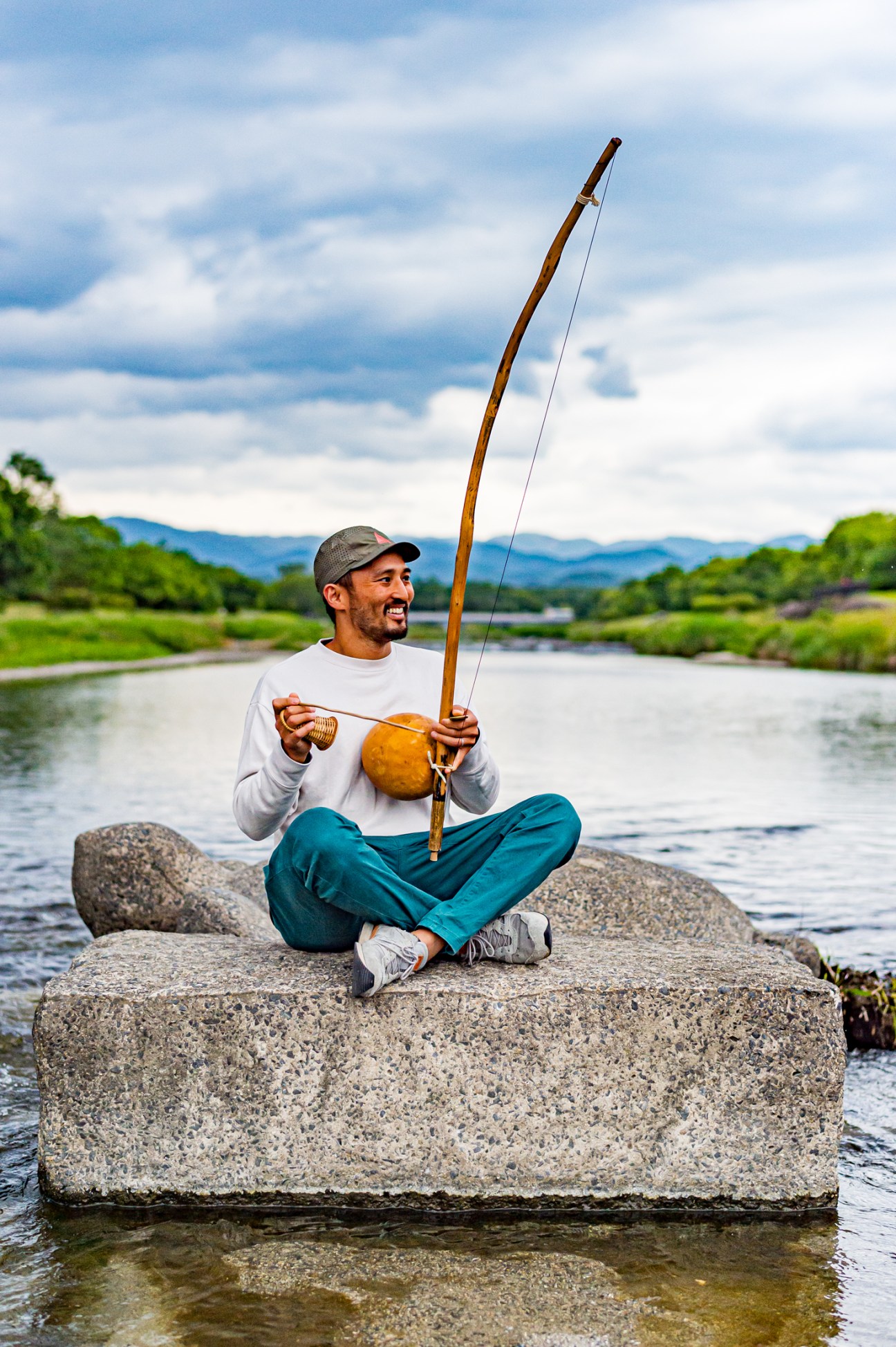 Man sitting cross-legged on stone block in river, holding traditional bow and gourd, mountains in background
