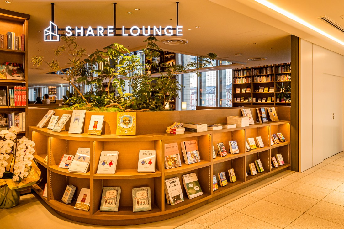 Curved wooden bookshelves displaying books and magazines in Share Lounge with plants and tall windows