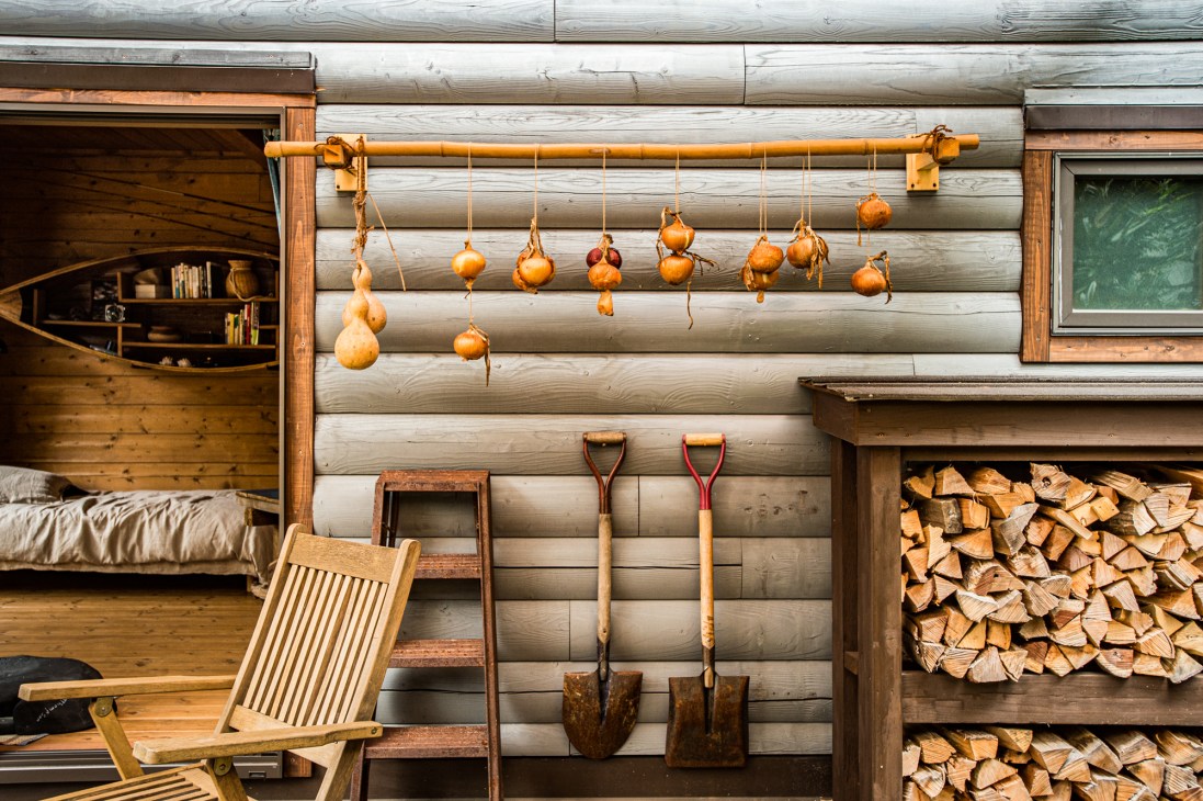 Exterior of a Bess log cabin with hanging gourds, shovels, firewood storage, and rustic furnishings against white-painted logs.