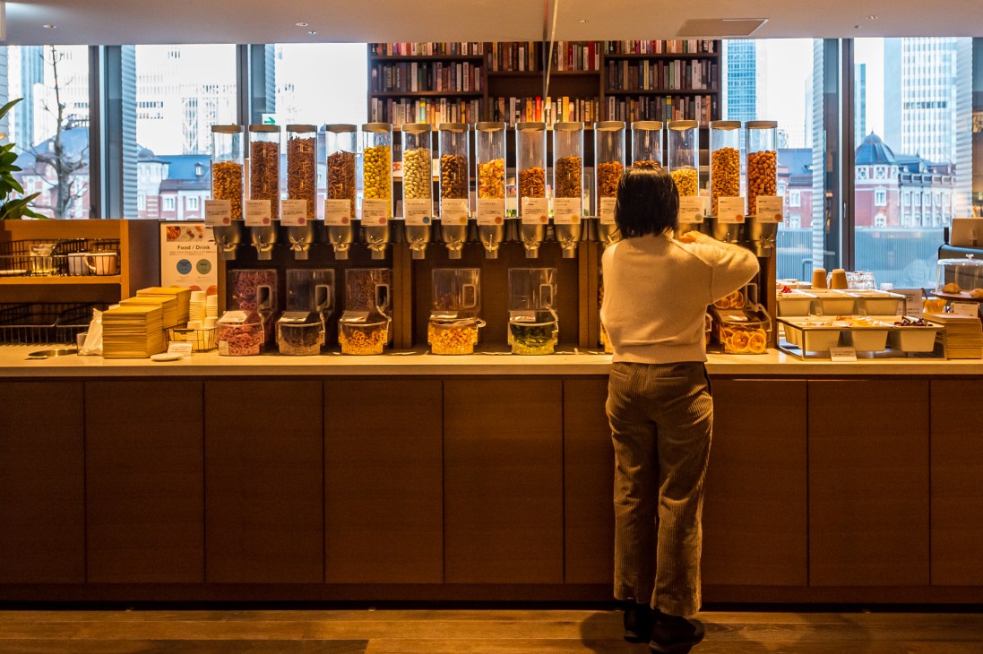 Person in beige sweater selecting items from wall-mounted dispensers containing nuts, cereals and snacks
