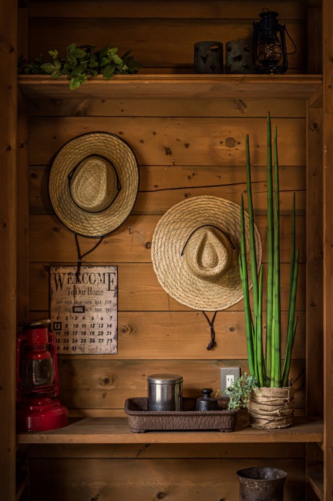 Wooden shelf displaying straw hats, potted plants, lantern and welcome calendar in rustic log home styling.