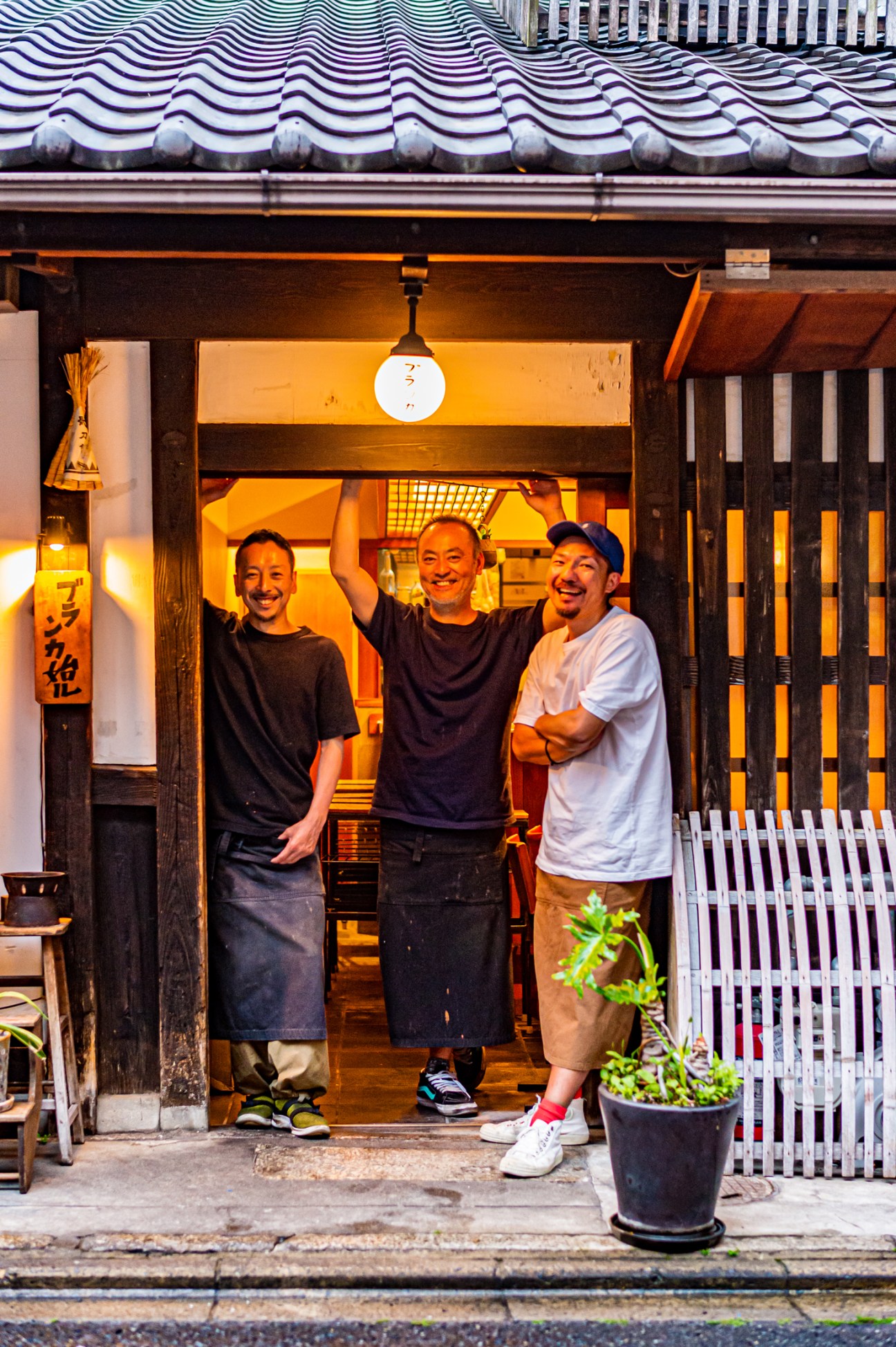 Three men smiling in the doorway of a traditional Japanese establishment with warm interior lighting, wooden facade, and decorative roof tiles