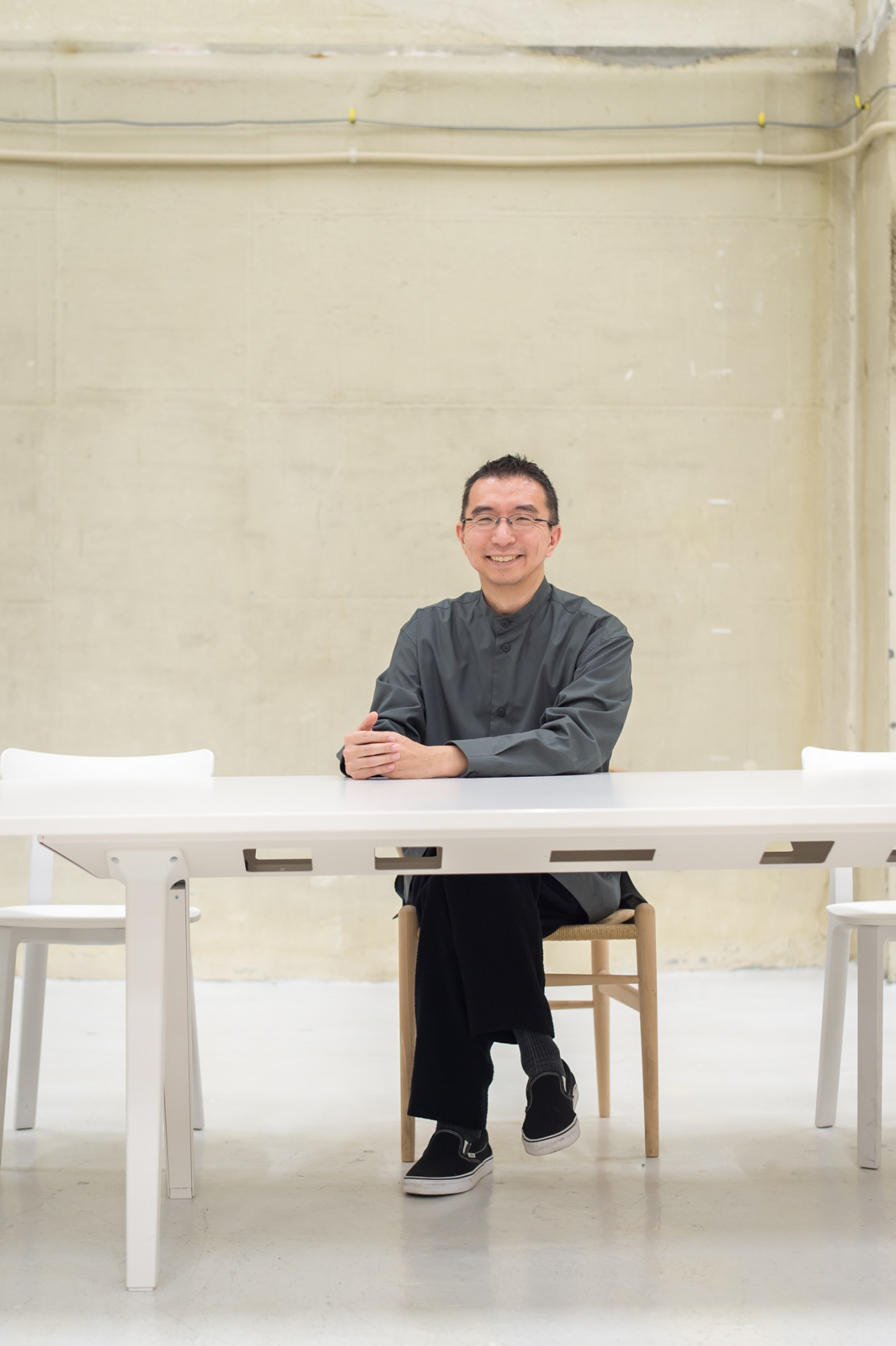 Sou Fujimoto sits smiling at a white table in a minimalist interior space wearing glasses and a grey jacket