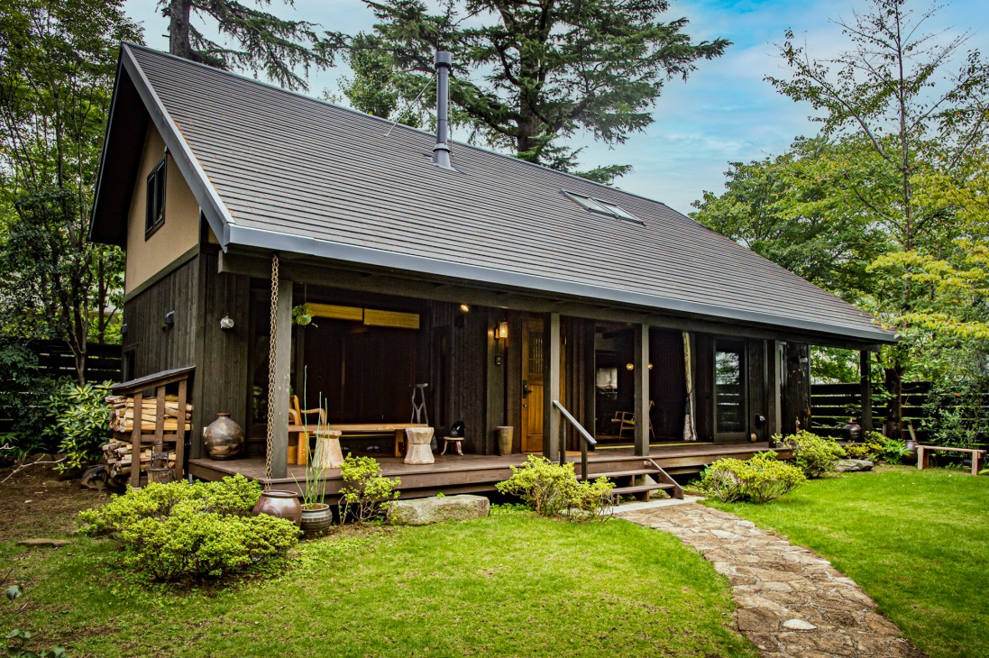 A modern Bess log cabin with black exterior, metal roof, and wooden terrace surrounded by green lawn and forest trees.