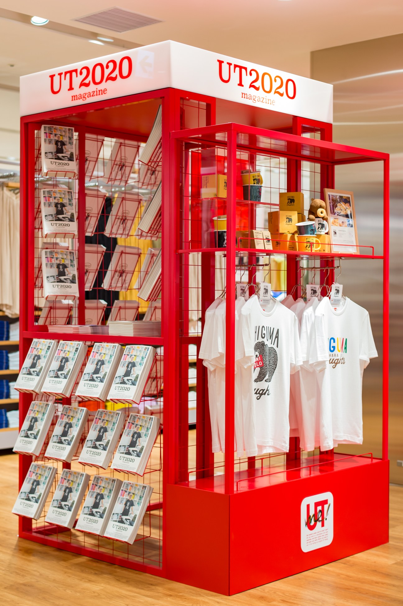 Red retail display kiosk for UT 2020 magazine with white t-shirts hanging inside and magazines on shelves.