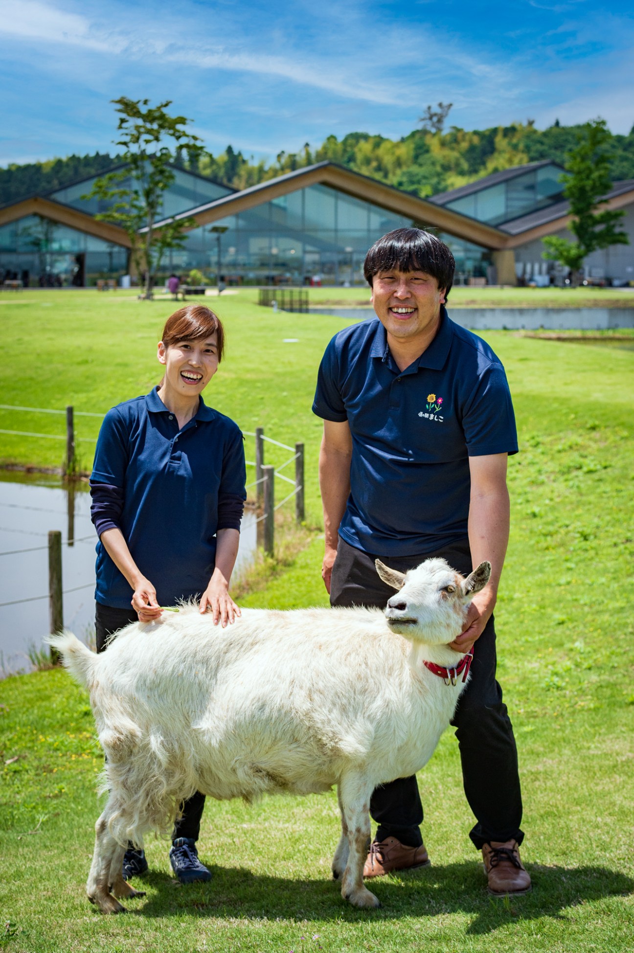 Two staff members in navy polo shirts pose with a white goat on a grass lawn in front of a modern building with large windows.