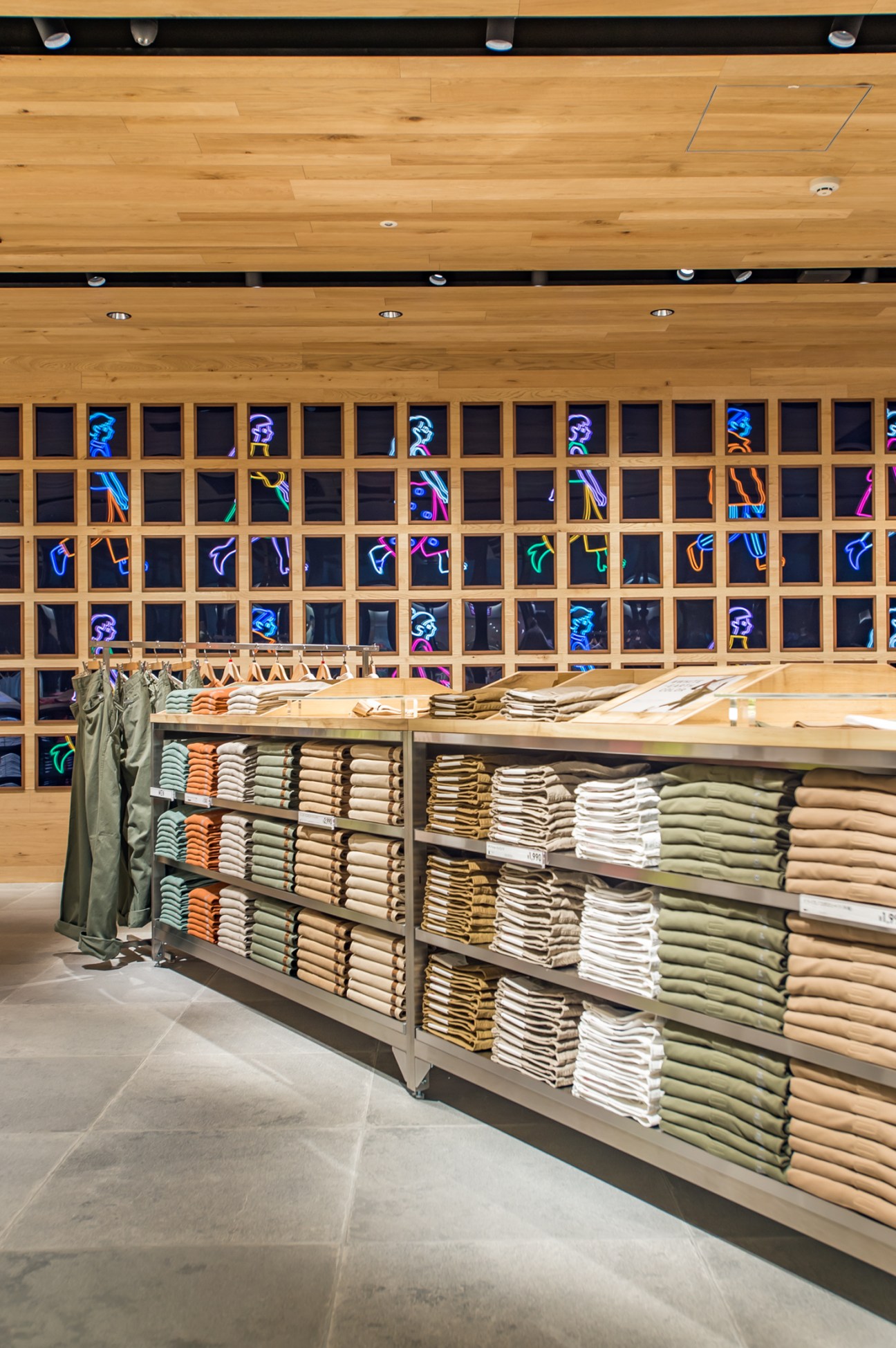 Folded T-shirts and apparel stacked neatly on display shelves with neon signage above at Uniqlo Harajuku store.