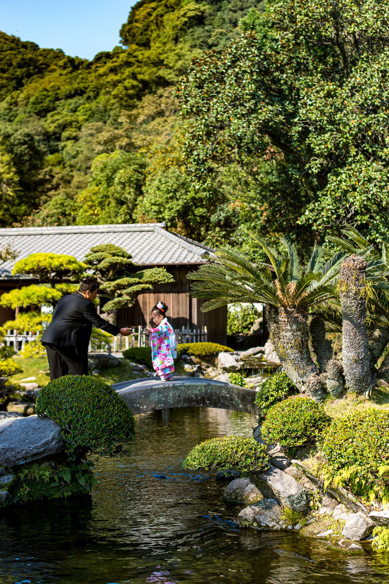 Man in black suit helping child in colorful kimono cross stone bridge over water in Japanese garden