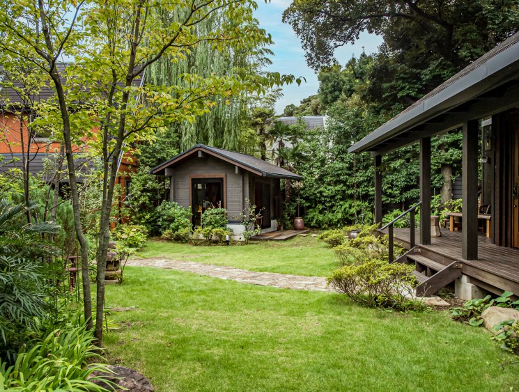 Wooden log cabin with extended deck surrounded by lush green trees and manicured lawn at Bess Magma Tokyo showroom.