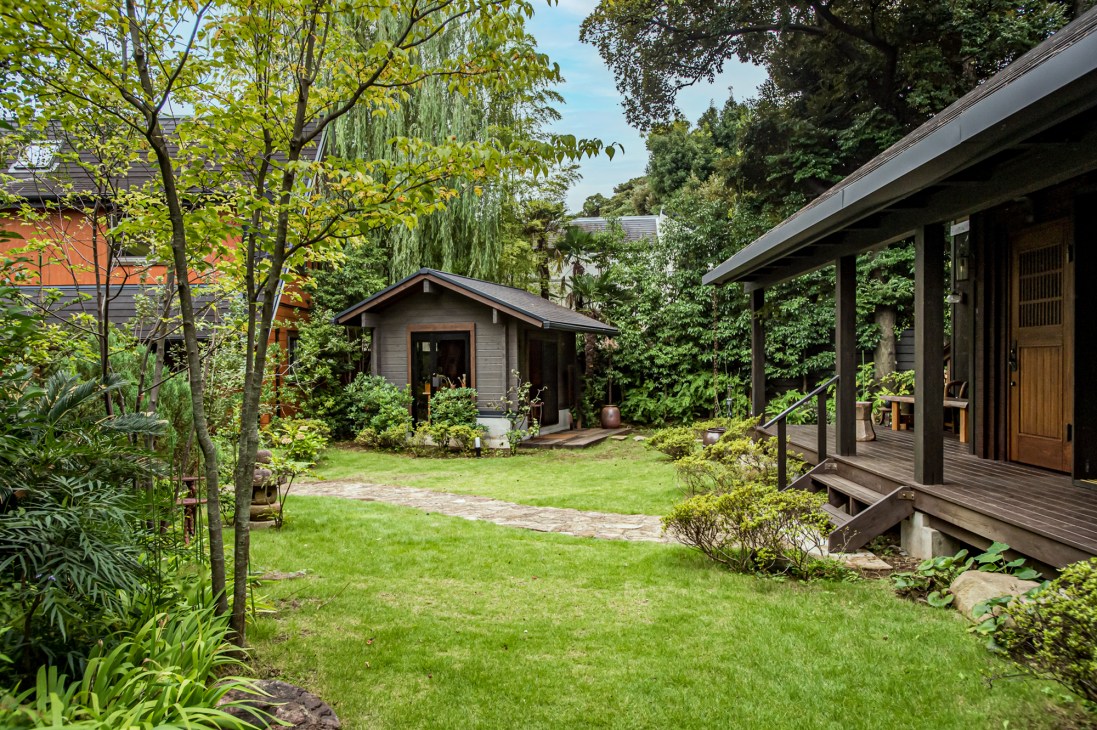 Wooden log cabin with extended deck surrounded by lush green trees and manicured lawn at Bess Magma Tokyo showroom.
