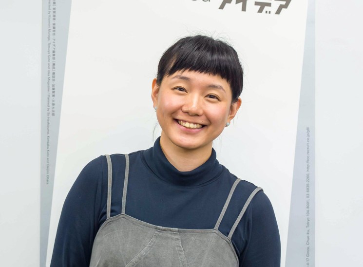 A smiling young woman with a short fringe wearing a navy turtleneck and dungarees against a white background.