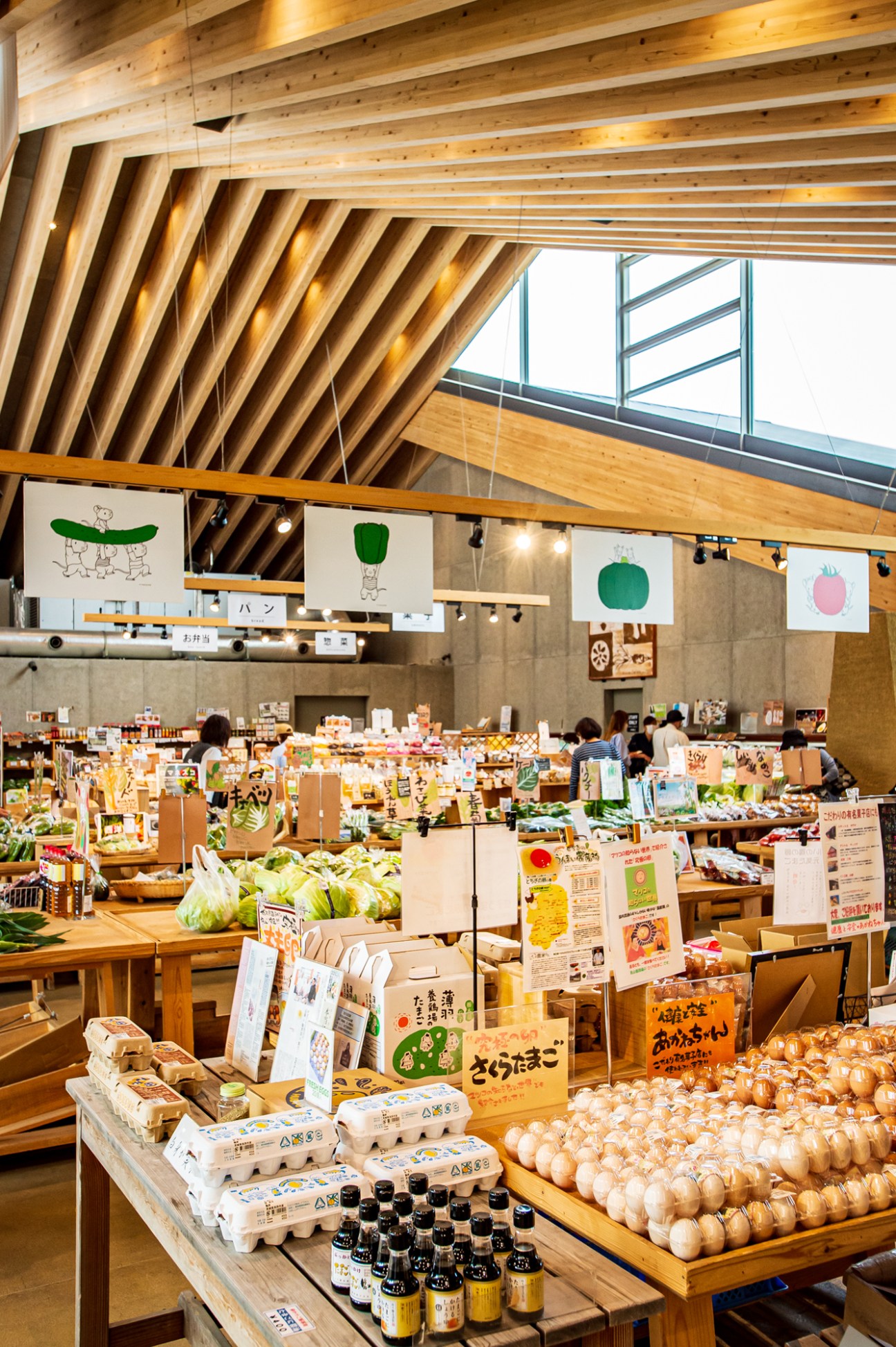 Interior of Mashiko Roadside Station with wooden ceiling beams, hanging illustrated signs, and tables displaying fresh farm produce including eggs, onions, and vegetables.